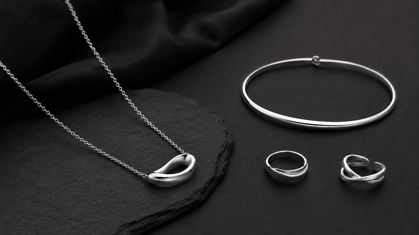 Silver necklace, bracelet and rings arranged on black surfaces